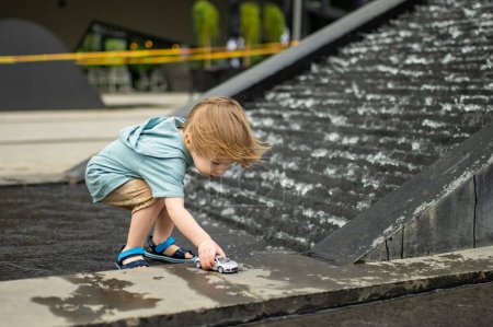 Téléchargez les photos : Mignon tout-petit garçon jouer avec jaune jouet voiture à l'extérieur. Enfant s'amuser avec de l'eau dans la fontaine de la ville. Activités estivales pour petits enfants. - en image libre de droit