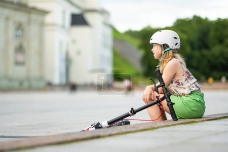Foto de Adorable jovencita montando su scooter en una ciudad en la soleada noche de verano. Niño bastante preadolescente montando un rodillo. Ocio activo y deportes al aire libre para niños. - Imagen libre de derechos