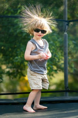 Téléchargez les photos : Joli tout-petit garçon avec des lunettes de soleil et des cheveux sales sautant sur un trampoline dans une arrière-cour. Sports et exercices pour les enfants. Activités de plein air estivales. - en image libre de droit