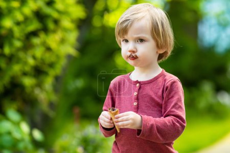 Téléchargez les photos : Mignon tout-petit garçon mangeant de la crème glacée fraîche savoureuse à l'extérieur par une chaude journée ensoleillée d'été. Les enfants mangent des bonbons. Aliments malsains pour enfants. - en image libre de droit