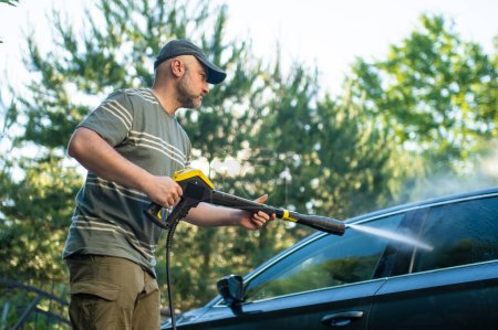 Téléchargez les photos : Young man using a water gun to wash his car. Male driver washing a car with contactless high pressure water jet. Cleaning a vehicle in a backyard. - en image libre de droit