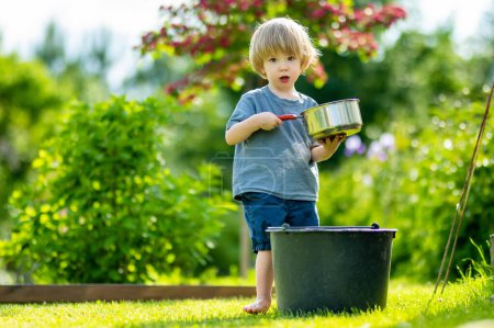 Photo for Cute blond little toddler playing with a cooking pot outdoors in the garden. Kid helping parents with gardening in the backyard in bright sunny summer day. - Royalty Free Image