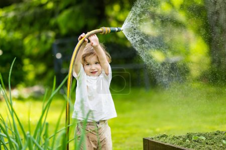 Foto de Cute toddler boy watering flower beds in the garden at summer day. Child using garden hose to water vegetables. Kid helping with everyday chores. Mommys little helper. - Imagen libre de derechos