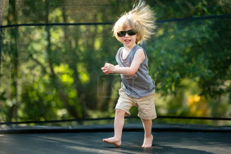 Photo for Cute toddler boy with sunglasses and messy hair jumping on a trampoline in a backyard. Sports and exercises for children. Summer outdoor leisure activities. - Royalty Free Image