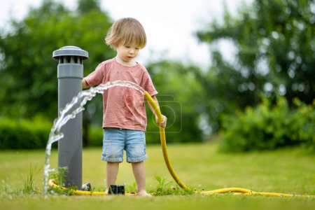 Téléchargez les photos : Cute toddler boy watering flower beds in the garden at summer day. Child using garden hose to water vegetables. Kid helping with everyday chores. Mommys little helper. - en image libre de droit