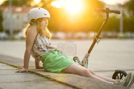 Photo for Adorable young girl riding her scooter in a city on sunny summer evening. Pretty preteen child riding a roller. Active leisure and outdoor sports for kids. - Royalty Free Image