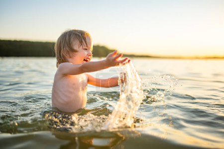 Foto de Lindo niño jugando junto a un lago en el caluroso día de verano. Adorable niño divirtiéndose al aire libre durante las vacaciones de verano. Actividades acuáticas para niños. - Imagen libre de derechos