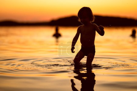 Téléchargez les photos : Petit garçon mignon jouant au bord d'un lac au coucher du soleil d'été. Adorable enfant s'amusant à l'extérieur pendant les vacances d'été. Activités aquatiques pour les enfants. - en image libre de droit