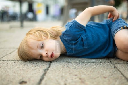 Foto de Niño pequeño teniendo un tamtro tendido en el suelo al aire libre. Maleducado niño en la calle. - Imagen libre de derechos