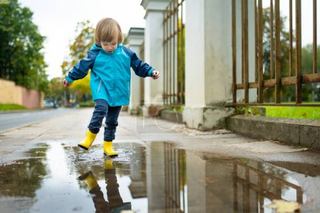 Photo for Adorable toddler boy wearing yellow rubber boots playing in a a puddle on sunny autumn day in city park. Child exploring nature. Fun autumn activities for small kids. - Royalty Free Image