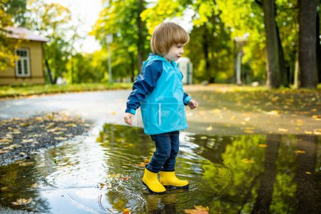 Adorable toddler boy wearing yellow rubber boots playing in a a puddle on sunny autumn day in city park. Child exploring nature. Fun autumn activities for small kids.