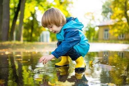Liebenswerter kleiner Junge in gelben Gummistiefeln, der an einem sonnigen Herbsttag im Stadtpark in einer Pfütze spielt. Kind erkundet die Natur. Spaßige Herbstaktivitäten für kleine Kinder.