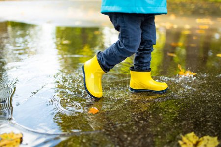 Liebenswerter kleiner Junge in gelben Gummistiefeln, der an einem sonnigen Herbsttag im Stadtpark in einer Pfütze spielt. Kind erkundet die Natur. Spaßige Herbstaktivitäten für kleine Kinder.