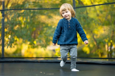 Téléchargez les photos : Petit garçon mignon sautant sur un trampoline dans une cour arrière par une journée d'été chaude et ensoleillée. Sports et exercices pour les enfants. Activités de plein air estivales. - en image libre de droit