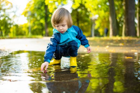 Adorable toddler boy wearing yellow rubber boots playing in a a puddle on sunny autumn day in city park. Child exploring nature. Fun autumn activities for small kids.