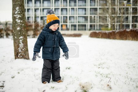 Téléchargez les photos : Adorable tout-petit garçon qui s'amuse dans une ville par une journée d'hiver enneigée. Mignon enfant portant des vêtements chauds jouant dans une neige. Activités hivernales pour les familles avec enfants. - en image libre de droit