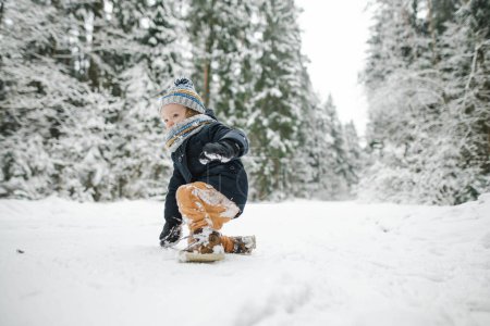 Foto de Lindo niño divirtiéndose en un paseo por el bosque de pinos cubierto de nieve en el frío día de invierno. Niño explorando la naturaleza. Actividades de invierno para niños pequeños. - Imagen libre de derechos