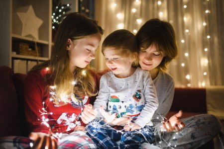Photo for Two big sisters and their toddler brother playing with Christmas lights in a cozy living room on Christmas eve. Kids spending time at home during winter break. Winter evening with family and kids. - Royalty Free Image
