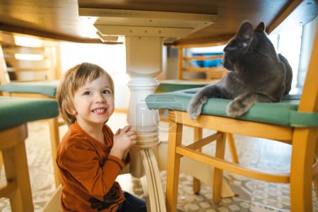 Foto de Lindo niño acariciando amistoso ruso azul pura raza gato debajo de la mesa. Divertido niño y su mascota en casa. - Imagen libre de derechos