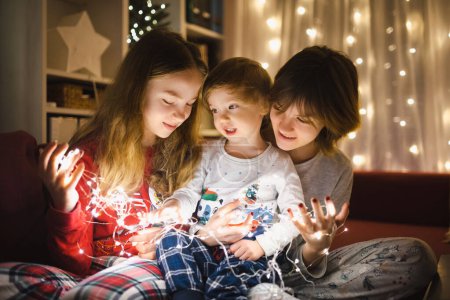 Téléchargez les photos : Deux grandes sœurs et leur petit frère jouant avec les lumières de Noël dans un salon confortable la veille de Noël. Les enfants passent du temps à la maison pendant les vacances d'hiver. Soirée d'hiver en famille et enfants. - en image libre de droit