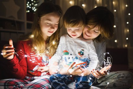 Photo for Two big sisters and their toddler brother playing with Christmas lights in a cozy living room on Christmas eve. Kids spending time at home during winter break. Winter evening with family and kids. - Royalty Free Image