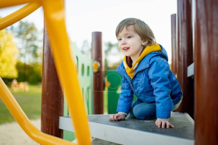 Téléchargez les photos : Petit garçon mignon s'amusant sur une aire de jeux à l'extérieur le jour chaud du printemps. Loisirs actifs pour les enfants au printemps. - en image libre de droit