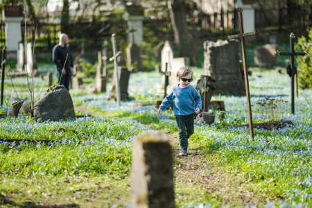 Netter kleiner Junge, der die blauen Frühlingsblumen der Scilla siberica bewundert, die im April auf dem Bernardiner Friedhof blühen, einem der drei ältesten Friedhöfe in Vilnius, Litauen.