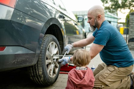 Photo for Cute toddler boy helping his father to change car wheels at their backyard. Father teaching his little son to use tools. Active parent of a small child. - Royalty Free Image