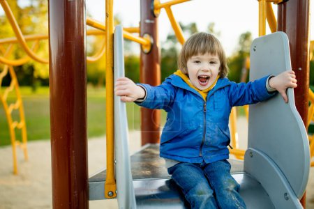 Téléchargez les photos : Petit garçon mignon s'amusant sur une aire de jeux à l'extérieur le jour chaud du printemps. Loisirs actifs pour les enfants au printemps. - en image libre de droit