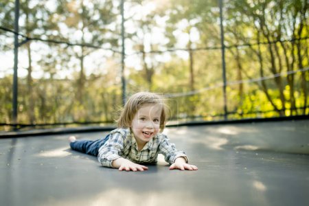 Téléchargez les photos : Joli petit garçon sautant sur un trampoline dans une cour par une chaude et ensoleillée journée d'été. Sports et exercices pour les enfants. Activités de plein air estivales. - en image libre de droit
