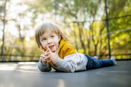 Foto de Lindo niño saltando en un trampolín en un patio trasero en el día de verano cálido y soleado. Deportes y ejercicios para niños. Actividades de ocio al aire libre de verano. - Imagen libre de derechos