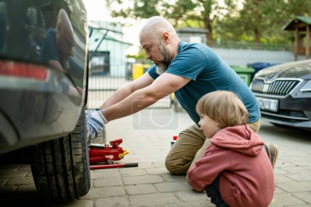 Téléchargez les photos : Petit garçon mignon aidant son père à changer de roues de voiture à leur arrière-cour. Père enseignant à son petit fils à utiliser des outils. Parents actifs d'un petit enfant. - en image libre de droit