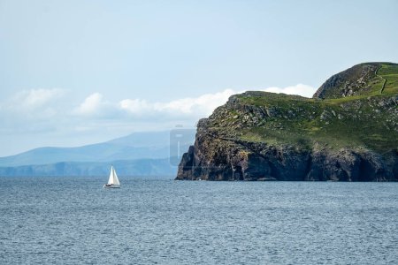 Foto de Pequeño velero flotando cerca de la costa áspera y rocosa a lo largo de la famosa ruta Ring of Kerry. Rugged coast of on Iveragh Peninsula, County Kerry, Irlanda. - Imagen libre de derechos