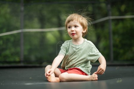 Téléchargez les photos : Joli petit garçon sautant sur un trampoline dans une cour par une chaude et ensoleillée journée d'été. Sports et exercices pour les enfants. Activités de plein air estivales. - en image libre de droit