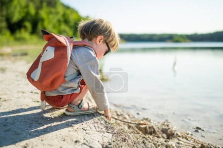 Téléchargez les photos : Joli petit garçon jouant au bord d'un lac ou d'une rivière par une chaude journée d'été. Adorable enfant s'amusant à l'extérieur pendant les vacances d'été. Activités aquatiques pour les enfants. - en image libre de droit