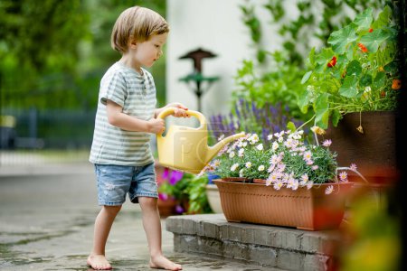 Téléchargez les photos : Mignon petit garçon arrosant les parterres de fleurs dans la cour arrière le jour d'été. Enfant utilisant l'arrosoir pour arroser les plantes. Un gamin qui aide dans les tâches quotidiennes. La petite assistante de maman. - en image libre de droit