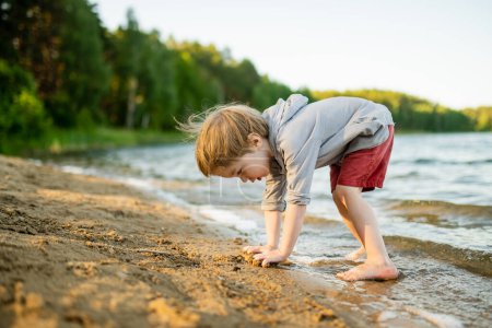 Foto de Lindo niño jugando junto a un lago o río en el caluroso día de verano. Adorable niño divirtiéndose al aire libre durante las vacaciones de verano. Actividades acuáticas para niños. - Imagen libre de derechos