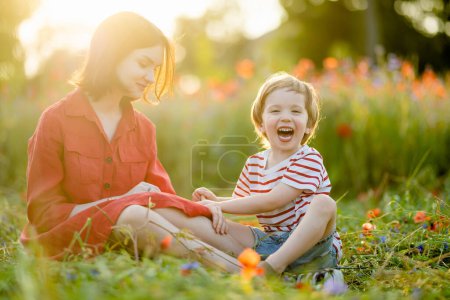 Photo for Beautiful teenage girl and her toddler brother admiring poppy and knapweed flowers in blossoming poppy field on sunny summer day. - Royalty Free Image