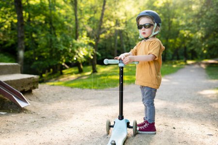 Téléchargez les photos : Adorable petit garçon chevauchant son scooter dans un parc de la ville lors d'une soirée d'été ensoleillée. Jeune enfant chevauchant un rouleau avec un casque. Loisirs actifs et sports de plein air pour enfants. - en image libre de droit