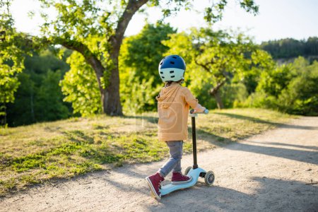 Photo for Adorable little boy riding his scooter in a city park on sunny summer evening. Young child riding a roller with a helmet on. Active leisure and outdoor sports for kids. - Royalty Free Image