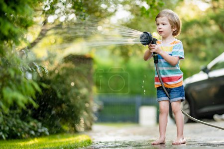 Téléchargez les photos : Mignon petit garçon jouant avec tuyau d'arrosage sur chaude journée d'été. Enfant jouant avec l'eau en été. Loisirs actifs avec les petits enfants. - en image libre de droit