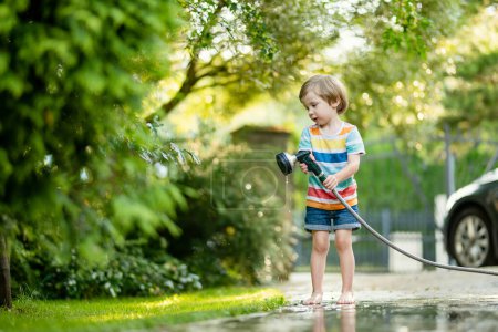 Téléchargez les photos : Mignon petit garçon jouant avec tuyau d'arrosage sur chaude journée d'été. Enfant jouant avec l'eau en été. Loisirs actifs avec les petits enfants. - en image libre de droit