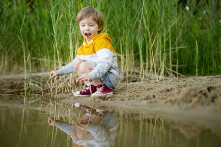 Foto de Lindo niño jugando junto a un lago o río en el caluroso día de verano. Adorable niño divirtiéndose al aire libre durante las vacaciones de verano. Actividades acuáticas para niños. - Imagen libre de derechos