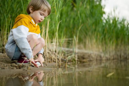 Téléchargez les photos : Joli petit garçon jouant au bord d'un lac ou d'une rivière par une chaude journée d'été. Adorable enfant s'amusant à l'extérieur pendant les vacances d'été. Activités aquatiques pour les enfants. - en image libre de droit