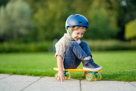 Téléchargez les photos : Joli petit garçon apprenant à faire du skateboard lors d'une belle journée d'été dans un parc. Enfant portant un casque de sécurité profitant d'une promenade en skateboard à l'extérieur. Loisirs actifs pour les petits enfants. - en image libre de droit