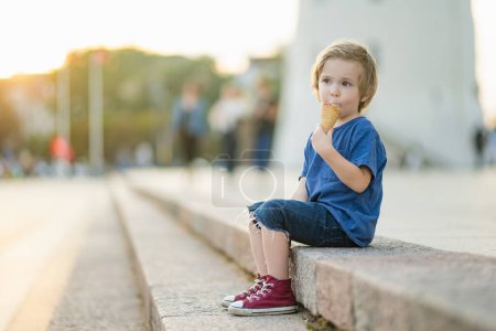 Foto de Lindo niño comiendo sabroso helado fresco al aire libre en el cálido día soleado de verano en Vilna, Lituania. Niño comiendo dulces. Alimento poco saludable para niños. - Imagen libre de derechos