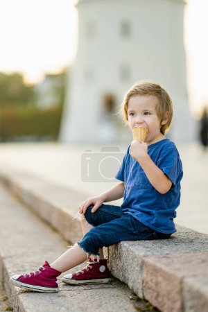 Foto de Cute little boy eating tasty fresh ice cream outdoors on warm sunny summer day in Vilnius, Lithuania. Child eating sweets. Unhealthy food for kids. - Imagen libre de derechos