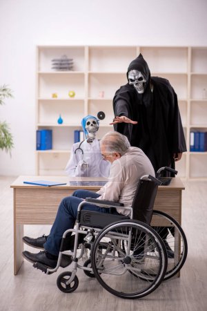 Photo for Old male patient in wheel-chair visiting two devil doctors - Royalty Free Image