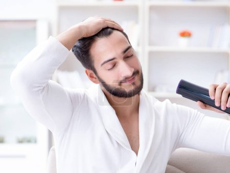 Téléchargez les photos : Le jeune homme sèche les cheveux à la maison avec un ventilateur sèche-cheveux - en image libre de droit
