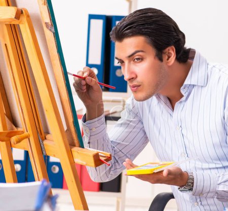 Foto de El joven empleado guapo disfrutando de la pintura en la oficina - Imagen libre de derechos
